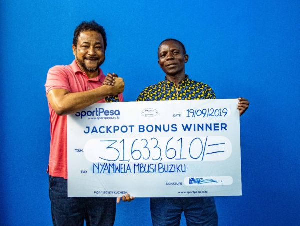 Mshindi wa BONUS ya SportPesa Jackpot, Nyamwela Mbusi akikabidhiwa mfano wa hundi ya Tsh 31,633,610 na Mkurugenzi wa Utawala an Udhibiti wa SportPesa Ndugu Tarimba Abbas